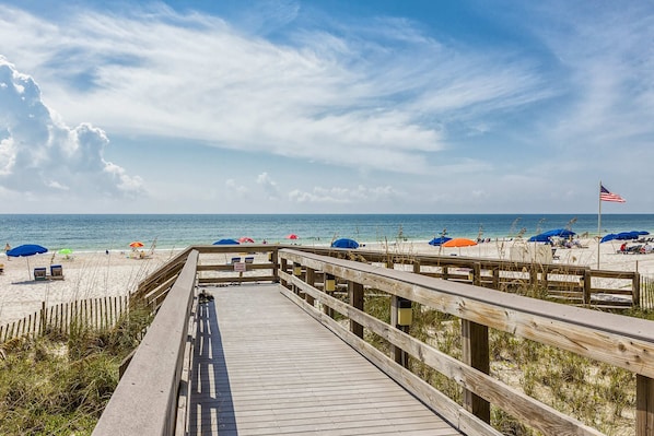 A wooden boardwalk leading to a beach with umbrellas and a flag. - Gulf-front condo with beach view, deck, AC, pools, hot tub & tennis (Orange Beach)