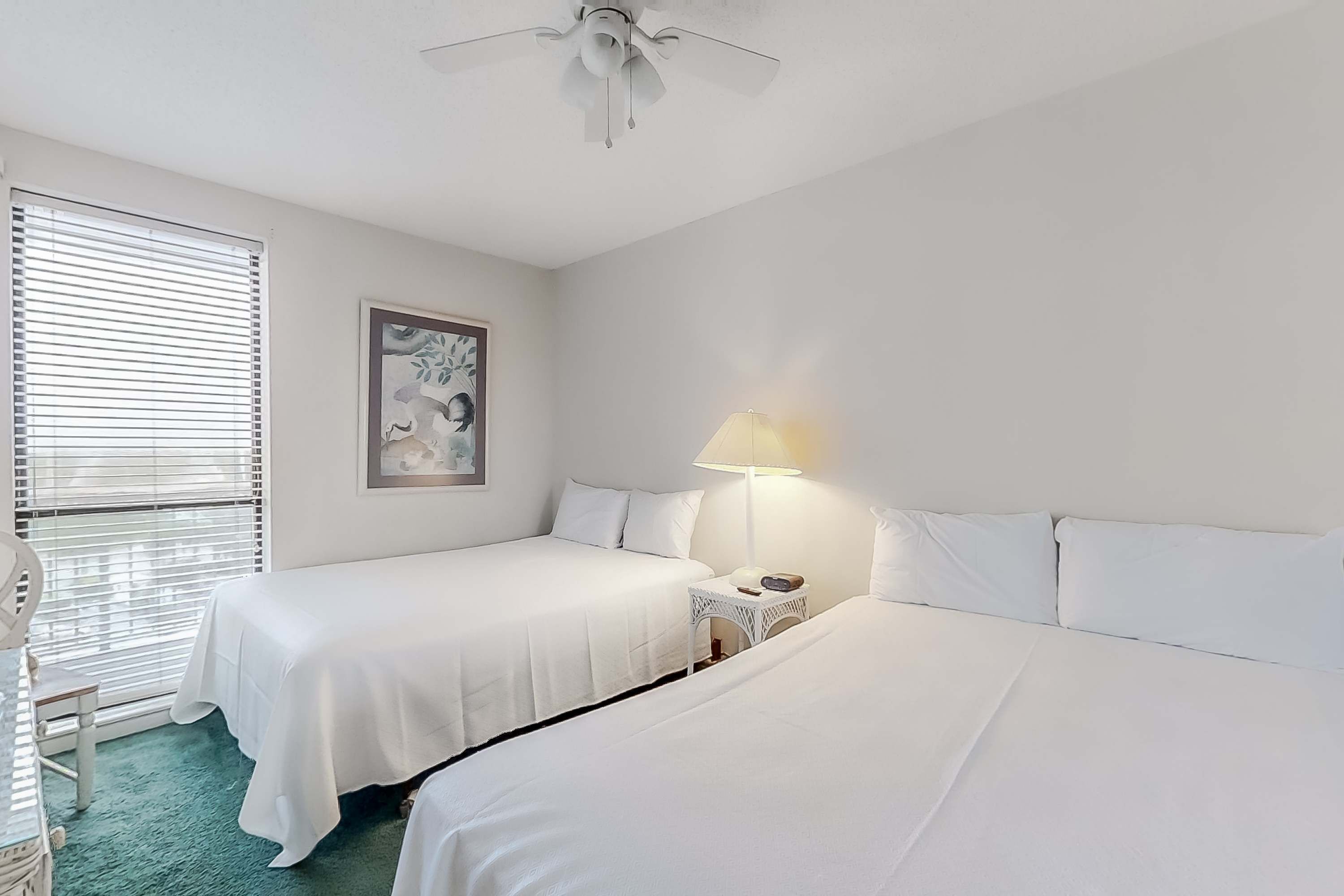 Habitación de hotel con dos camas, ventilador de techo, lámpara y una obra de arte enmarcada en la pared.