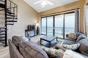 A living room with a sofa, coffee table, TV, and a balcony with a view. - Gulf-front corner condo with wraparound balcony, pools, beach access (Orange Beach)