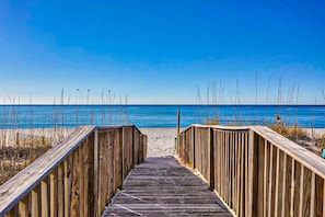 Ein hölzerner Steg führt zu einem Strand bei blauem Himmel.