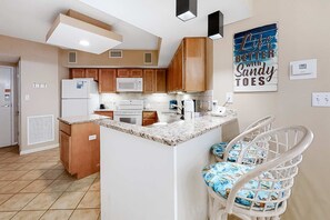 Une cuisine avec des meubles en bois, un réfrigérateur blanc, un four à micro-ondes et une machine à café. On y trouve également un coin bar avec un tabouret blanc et un coussin bleu.