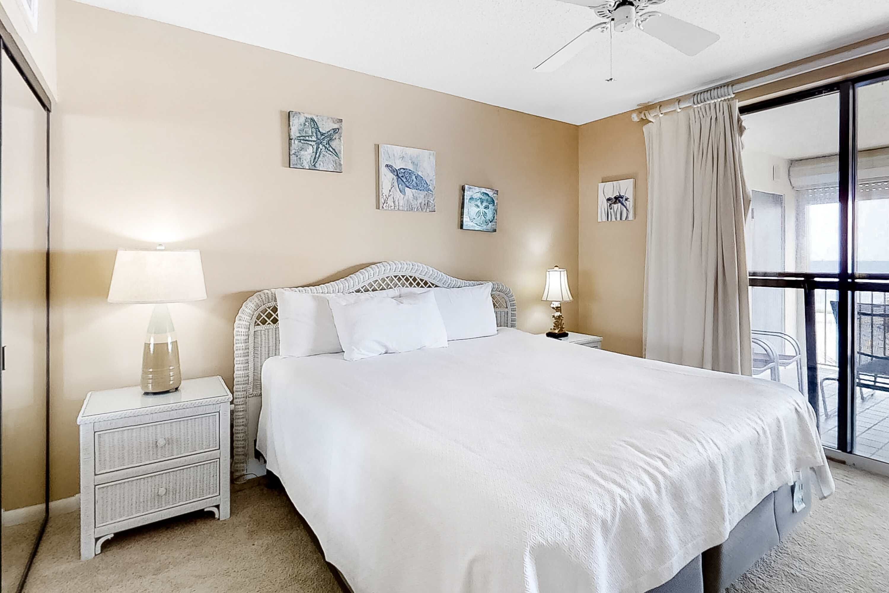 Une chambre d’hôtel avec un lit, deux tables de chevet, un ventilateur de plafond, des œuvres d’art accrochées aux murs et une grande fenêtre avec des rideaux.