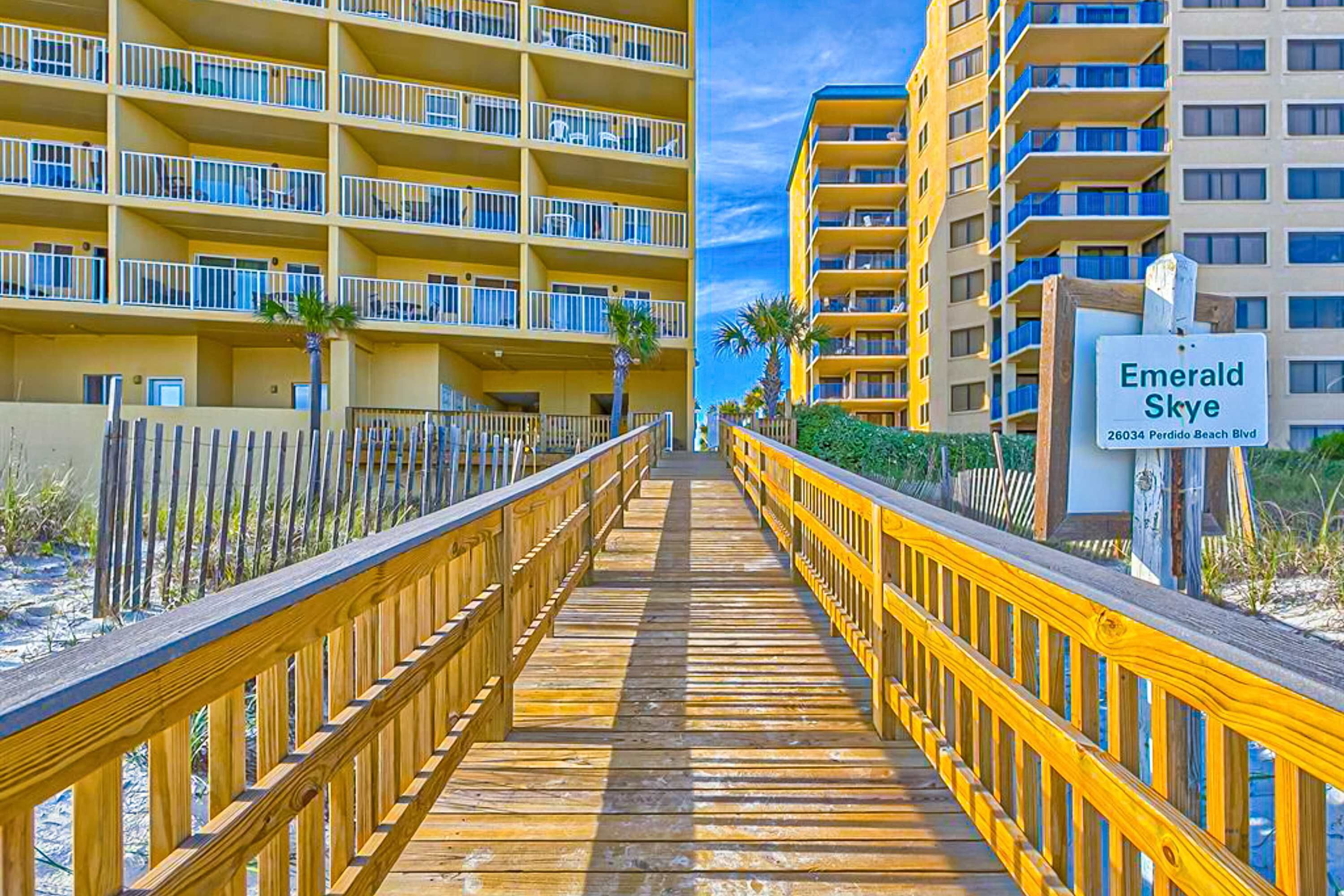 Un muelle de madera que conduce a un hotel con múltiples balcones y un letrero que dice 'Emerald Skye 26034 Perdido Beach Blvd'.