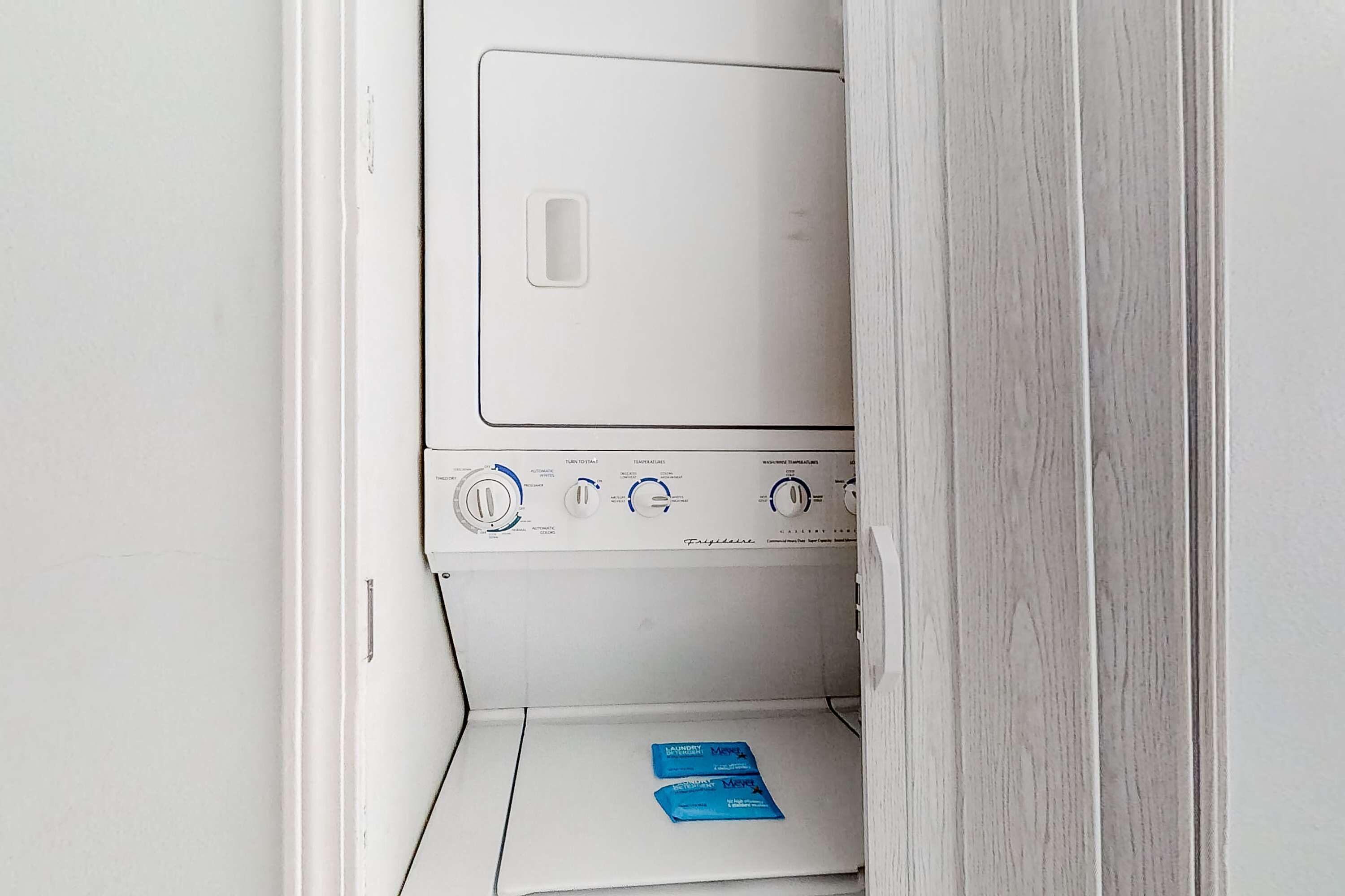 A white laundry room with a front-loading washer and dryer, a control panel with various settings, and two blue laundry detergent packets on the floor.