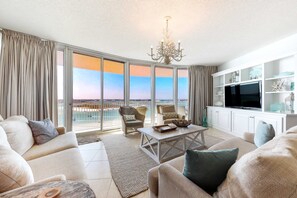 A spacious living room with a large window, a chandelier, and a flat-screen TV. - Gulf-front 8th-floor Condo with balcony, pool, hot tubs, & tennis courts (Orange Beach)