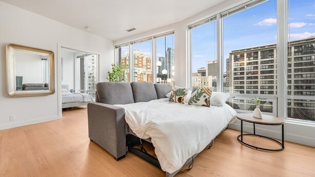 A modern bedroom with a bed, a grey sofa, a small table, and large windows offering a city view. - Modern & Cozy Stay in Prime DT (Vancouver)