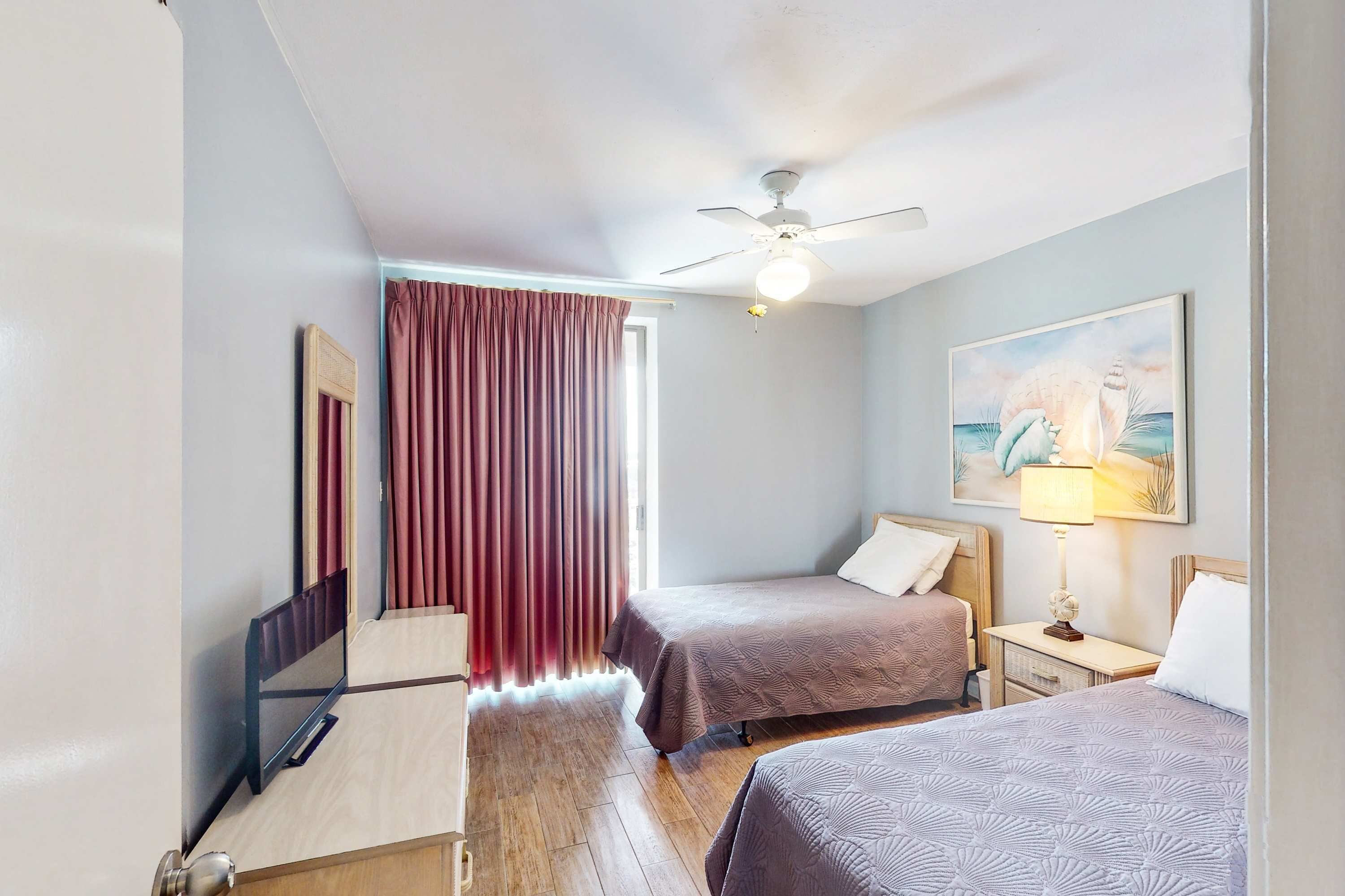 A hotel room with two beds, a ceiling fan, a painting, a lamp, and a window with curtains.