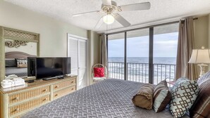Ein Schlafzimmer mit einem großen Bett, einem Deckenventilator, einem Fernseher auf einer Kommode, einem Sessel und Blick aufs Meer.