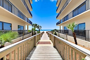 A wooden pathway leading to a beach with a clear sky, surrounded by buildings with balconies. - Beach front paradise w/ beautiful views, outdoor pool, & sauna (Orange Beach)