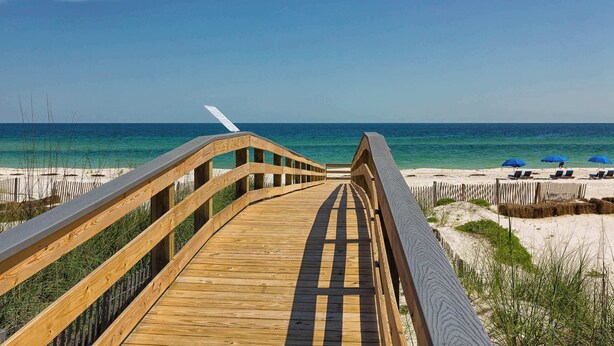 Een houten looppad dat naar een strand leidt, met helderblauw water en enkele parasols.