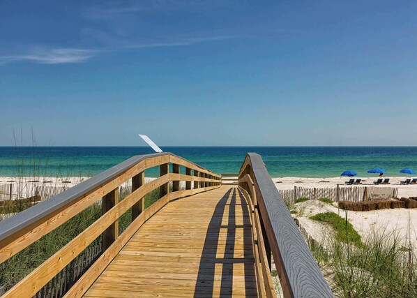 A wooden boardwalk leading to a beach with clear blue water and a few umbrellas. - Beach view beach home w community outdoor pool, sun deck & private beach (Orange Beach)
