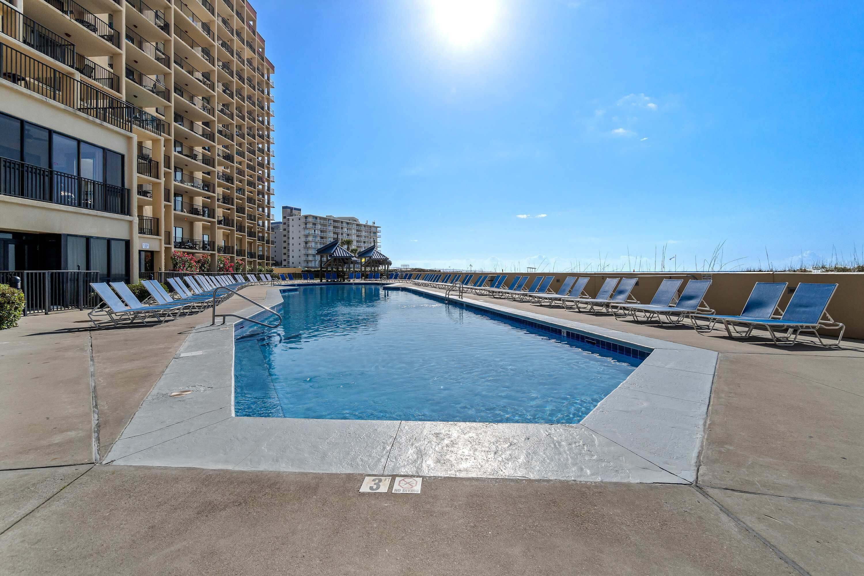 A swimming pool with lounge chairs, a building with balconies, and a clear sky.