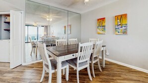 Een eetgedeelte met een houten tafel en witte stoelen, een plafondventilator en twee schilderijen aan de muur.