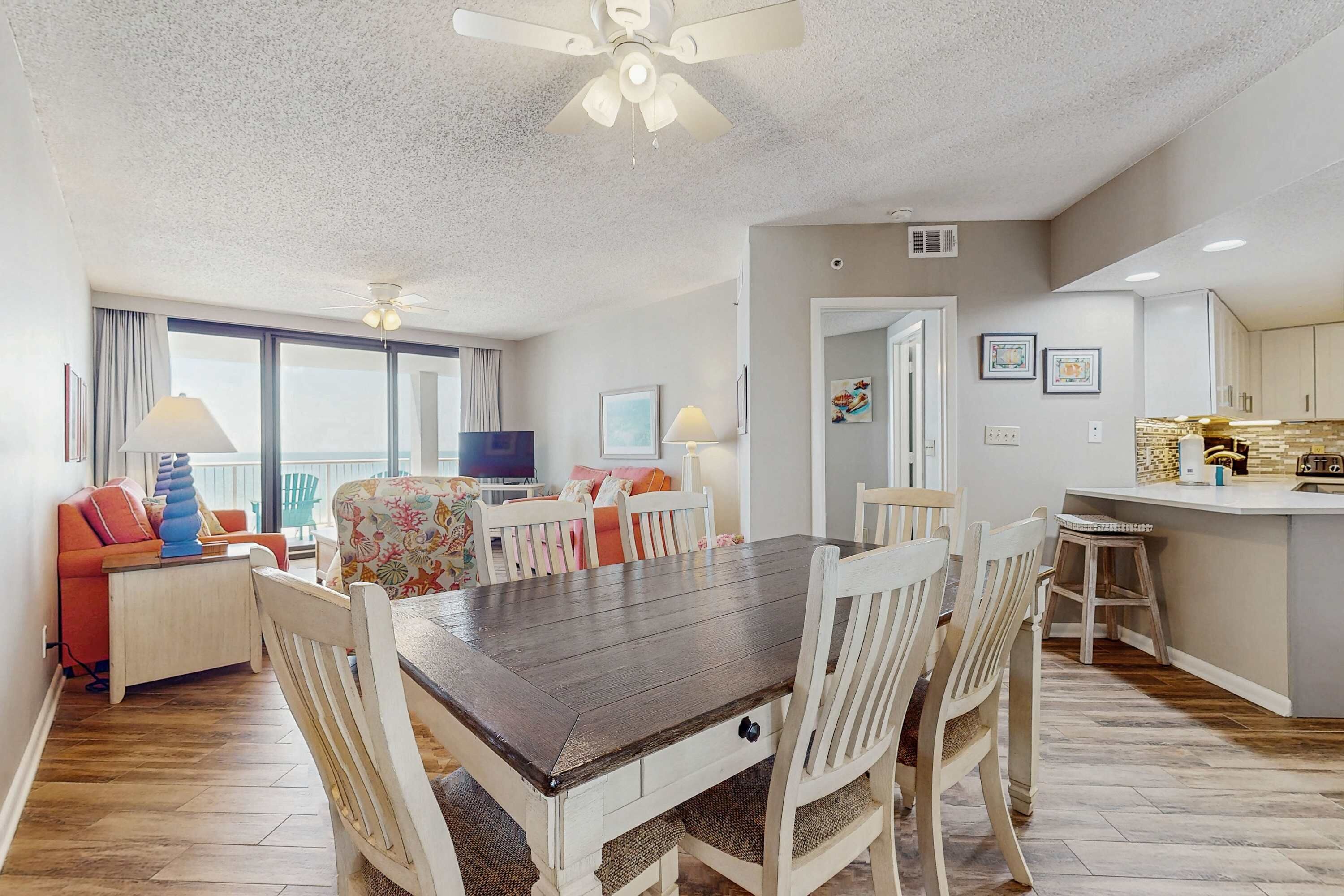 Un coin repas avec une table en bois et des chaises blanches, une cuisine avec un bar, et un salon avec un canapé et une télévision.