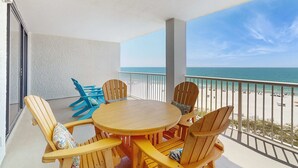 Een balkon met een houten tafel en stoelen, uitzicht op het strand, en een hekwerk.