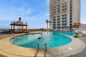 A swimming pool with a gazebo, lounge chairs, and a tall building in the background. - Elegant beachfront condo with wraparound balcony, Gulf views & pools (Orange Beach)