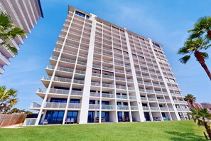 A multi-story hotel with balconies, a flat roof, and a grassy area in front. - Elegant beachfront condo with wraparound balcony, Gulf views & pools (Orange Beach)