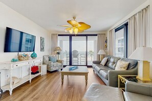 A modern living room with a flat-screen TV, a ceiling fan, a sofa, a coffee table, and a balcony with a view. - Elegant beachfront condo with wraparound balcony, Gulf views & pools (Orange Beach)