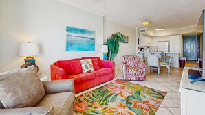 A living room with a red sofa, a floral rug, a ceiling fan, and a kitchen in the background. - Beachfront property with Gulf-view balcony, outdoor pool & hot tub - near shops (Orange Beach)