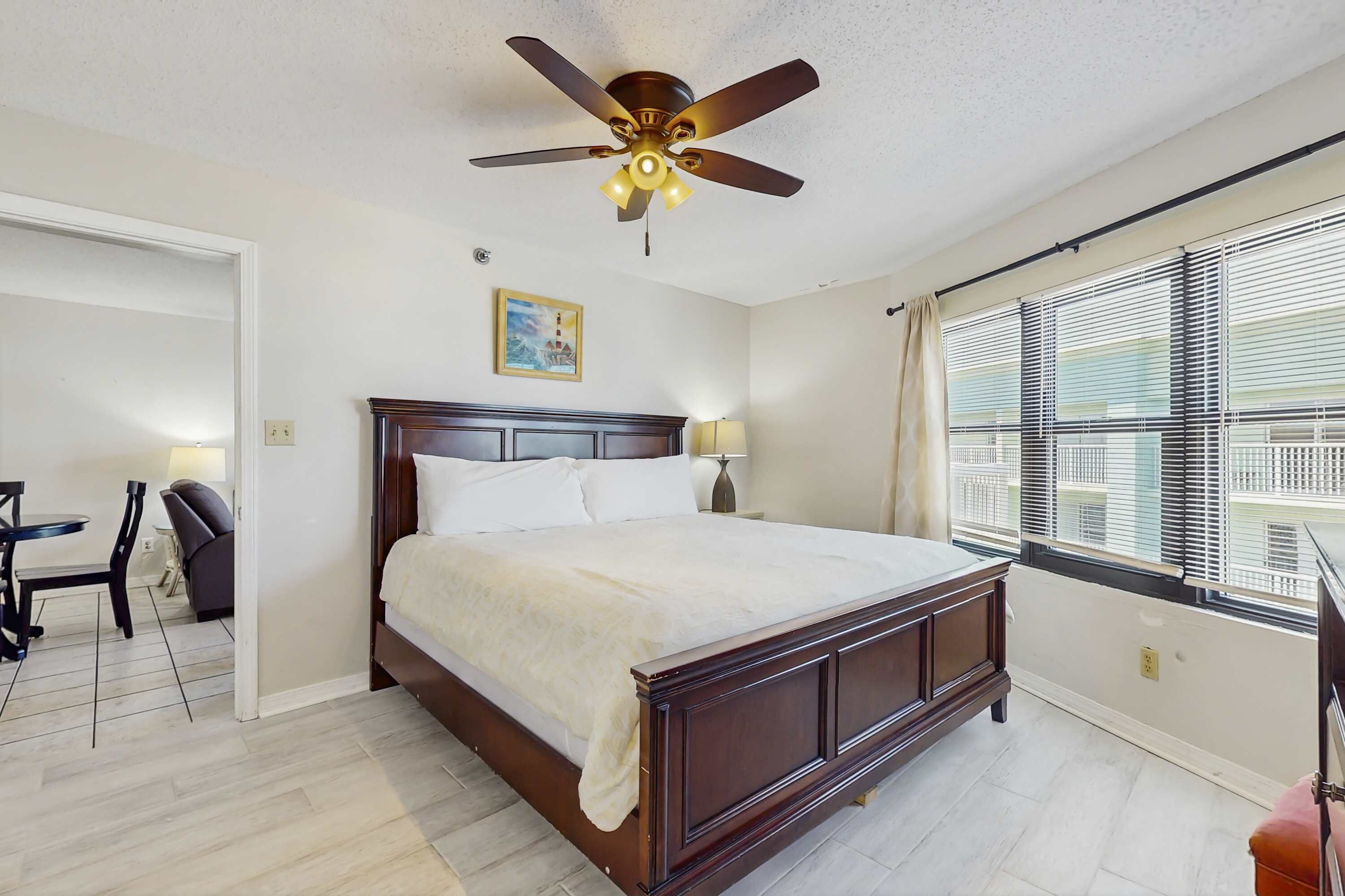 Une chambre à coucher comprenant un lit, un ventilateur de plafond, un bureau avec une chaise et une fenêtre avec des stores.