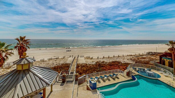 A beach view with a pool area, including lounge chairs and a small building. - Spacious condo with Gulf-front views & access to pools, hot tub, gym (Orange Beach)