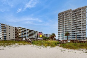 A beachfront hotel with multiple balconies, a sandy beach, and a flagpole with flags. - Beachfront condo with Gulf views, beach access, in/outdoor pools, elevator, W/D (Orange Beach)