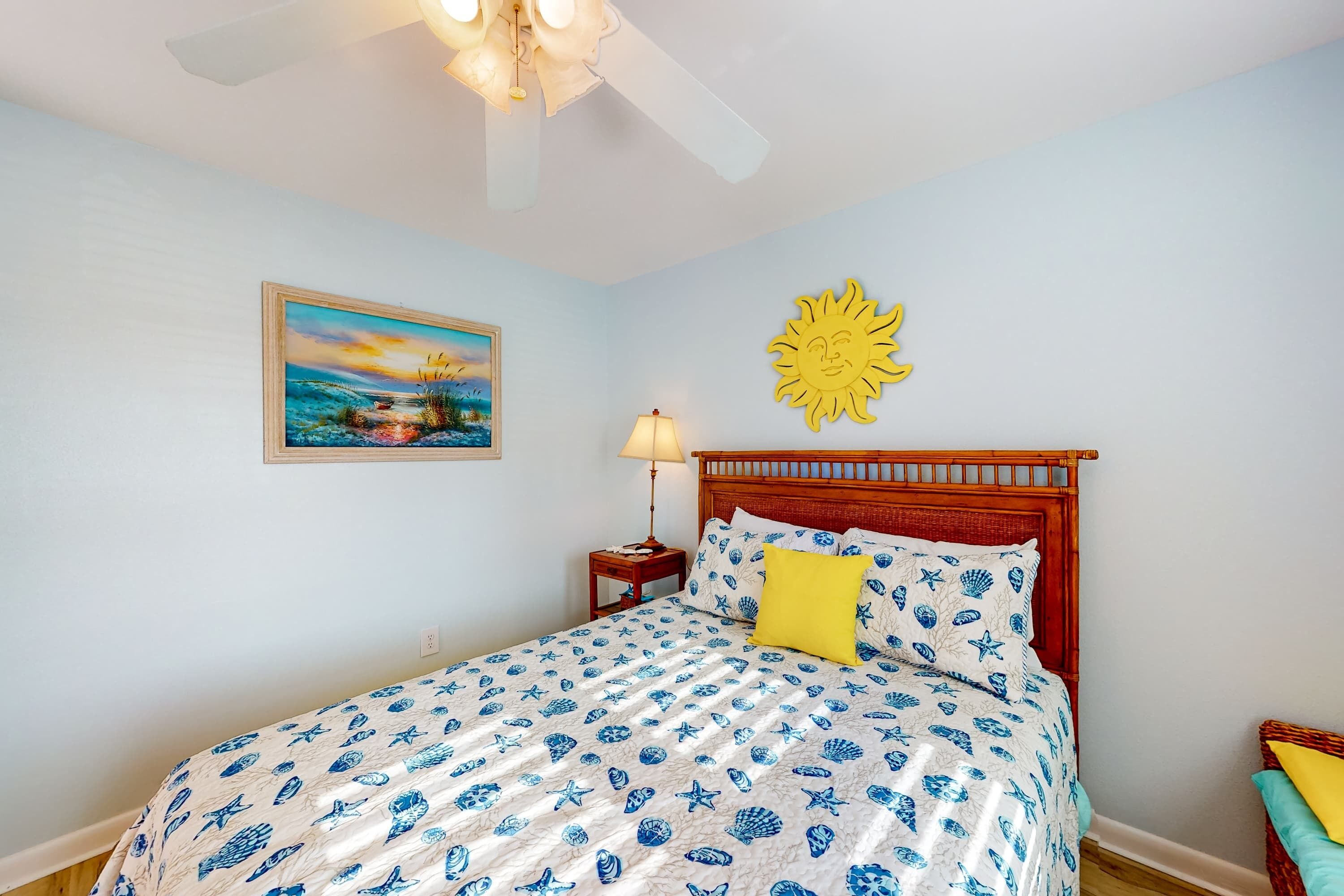 A bedroom with a bed, a nightstand, a lamp, a ceiling fan, a painting, and a sun decoration.