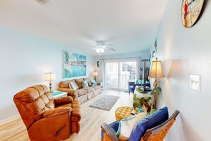 A living room with a sofa, armchair, and a coffee table. There is a ceiling fan, a painting on the wall, and a window with curtains.