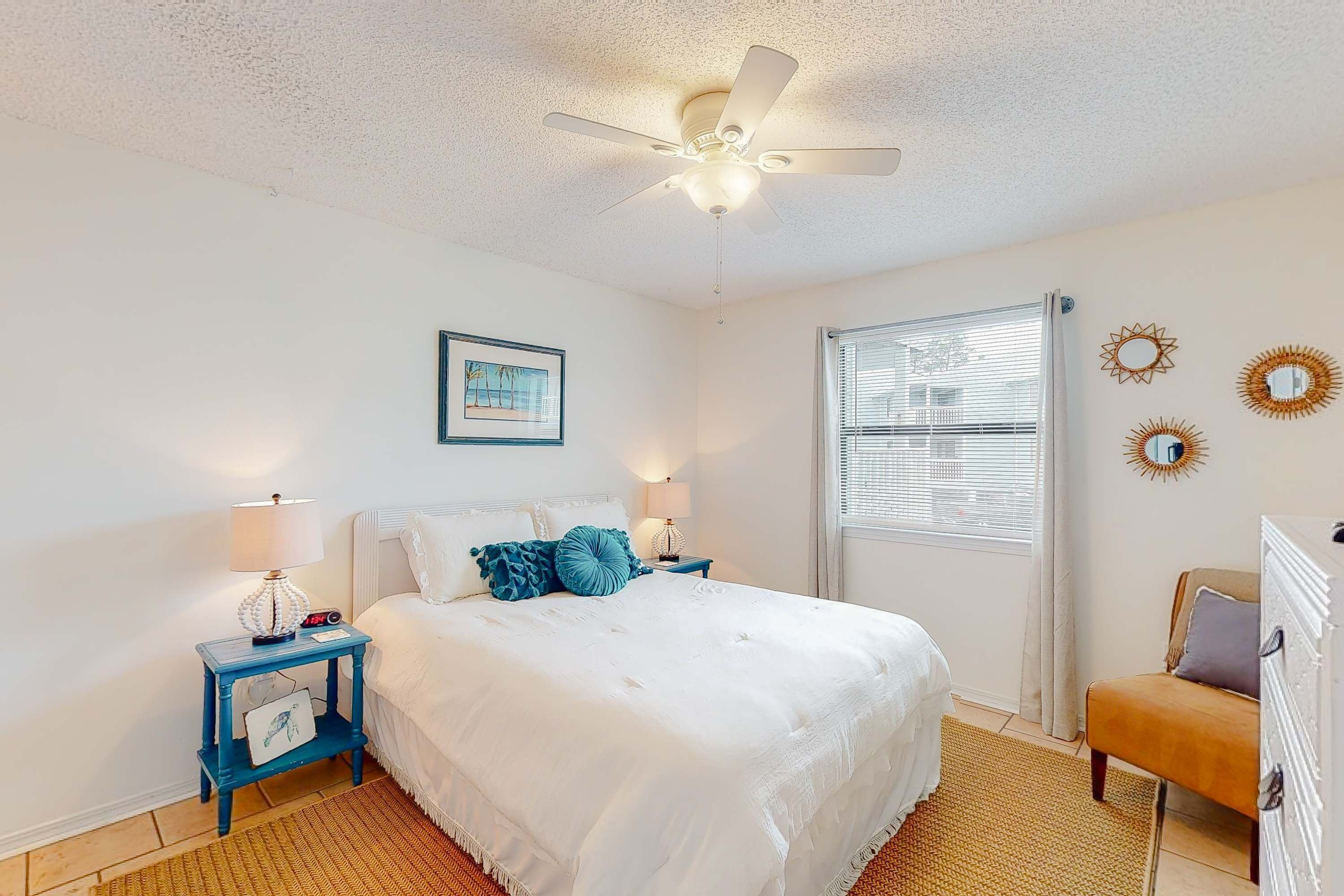 Un dormitorio con cama, mesita de noche, lámpara, ventilador de techo, ventana y espejos decorativos en la pared.