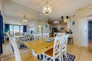 A modern living room with a wooden dining table, white chairs, and a hanging light fixture. - Beachfront 10th-floor condo with balcony, pools, hot tub, gym, WiFi, & AC (Orange Beach)