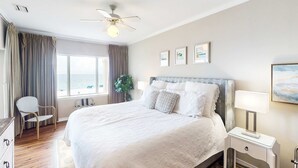 Ein Hotelzimmer mit Bett, Deckenventilator, Fenster mit Vorhängen, Nachttisch mit Lampe und gerahmten Bildern an der Wand.