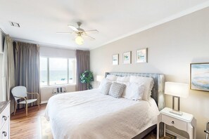 A hotel room with a bed, a ceiling fan, a window with curtains, a bedside table with a lamp, and framed artwork on the wall. - Beachfront at Marlin Key, Dazzling Views, Pool & Private Boardwalk to Sand (Orange Beach)