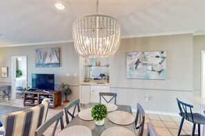A dining area with a chandelier, a TV, and artwork on the walls. - Beachfront at Marlin Key, Dazzling Views, Pool & Private Boardwalk to Sand (Orange Beach)