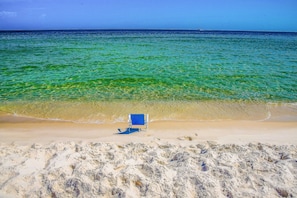 A beach with clear turquoise water, a sandy shore, and a blue beach chair. - White Caps | Beachfront | Pools, Hot Tub & Tennis | Gulf-View Balcony (Orange Beach)