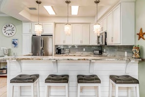A kitchen with white cabinets, a stainless steel refrigerator, a marble countertop, and three bar stools. - Ground-floor condo next to on-site pool with private patio & beach access (Dauphin Island)