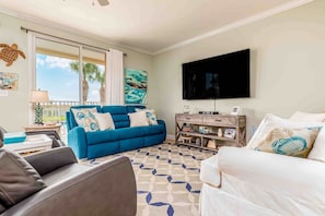 A living room with a blue sofa, a white sofa, a flat-screen TV, and a patterned rug. - Ground-floor condo next to on-site pool with private patio & beach access (Dauphin Island)