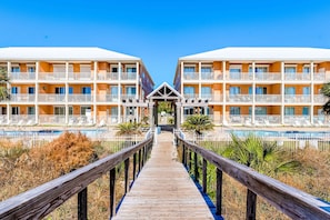 A hotel with a wooden walkway leading to a pool area, surrounded by palm trees and a clear blue sky. - Ground-floor condo next to on-site pool with private patio & beach access (Dauphin Island)