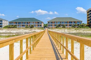 Ein Holzsteg führt zu Gebäuden direkt am Strand, mit Balkonen und wolkenlosem Himmel.