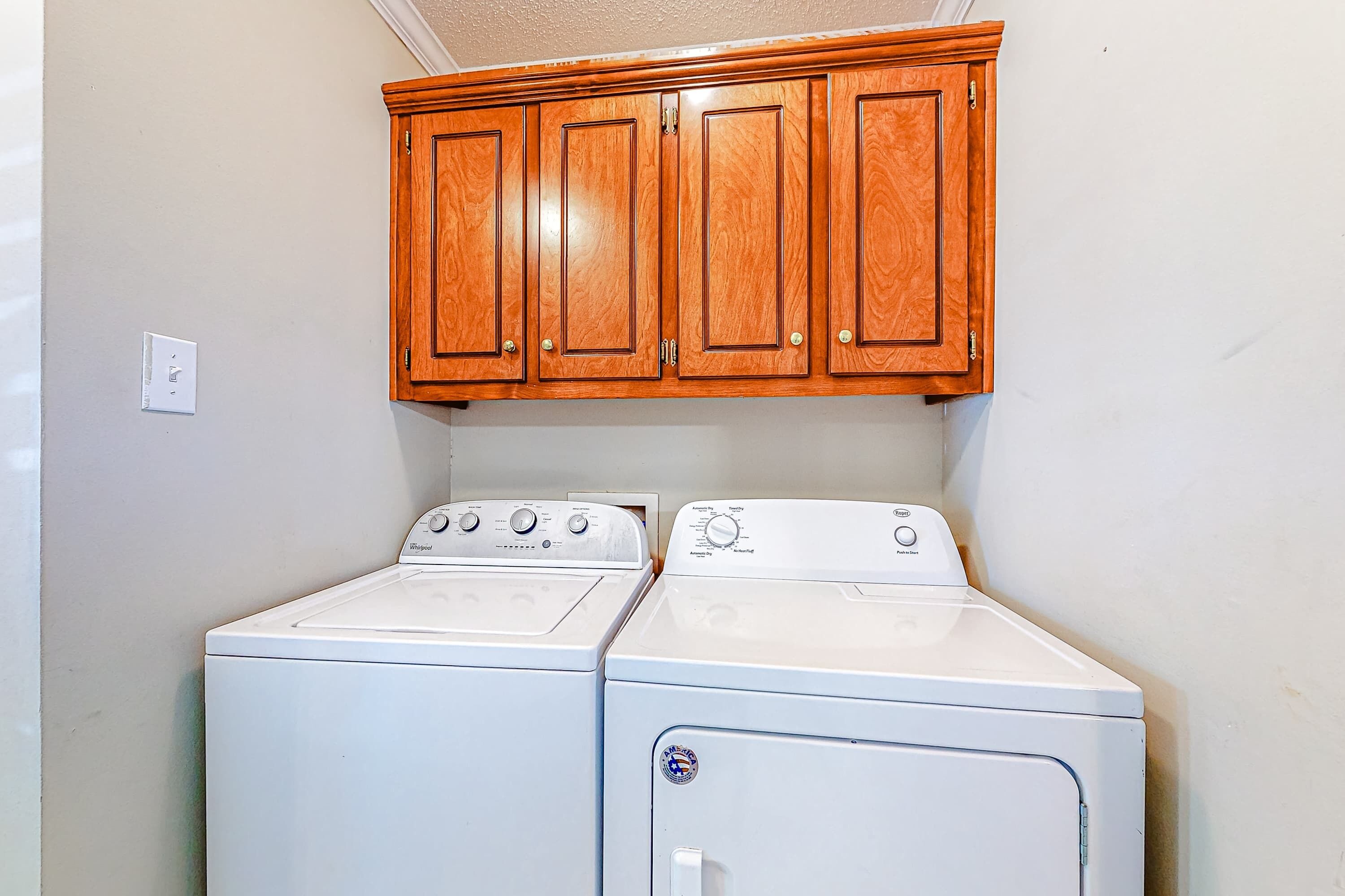 A laundry room with two white front-loading washers and dryers, wooden cabinets above, and a light switch on the wall.