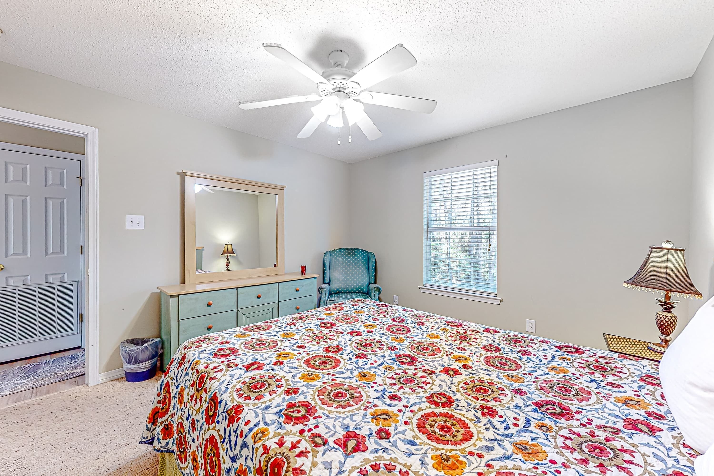 A bedroom with a bed, a ceiling fan, a window with blinds, a chair, a dresser, and a lamp.