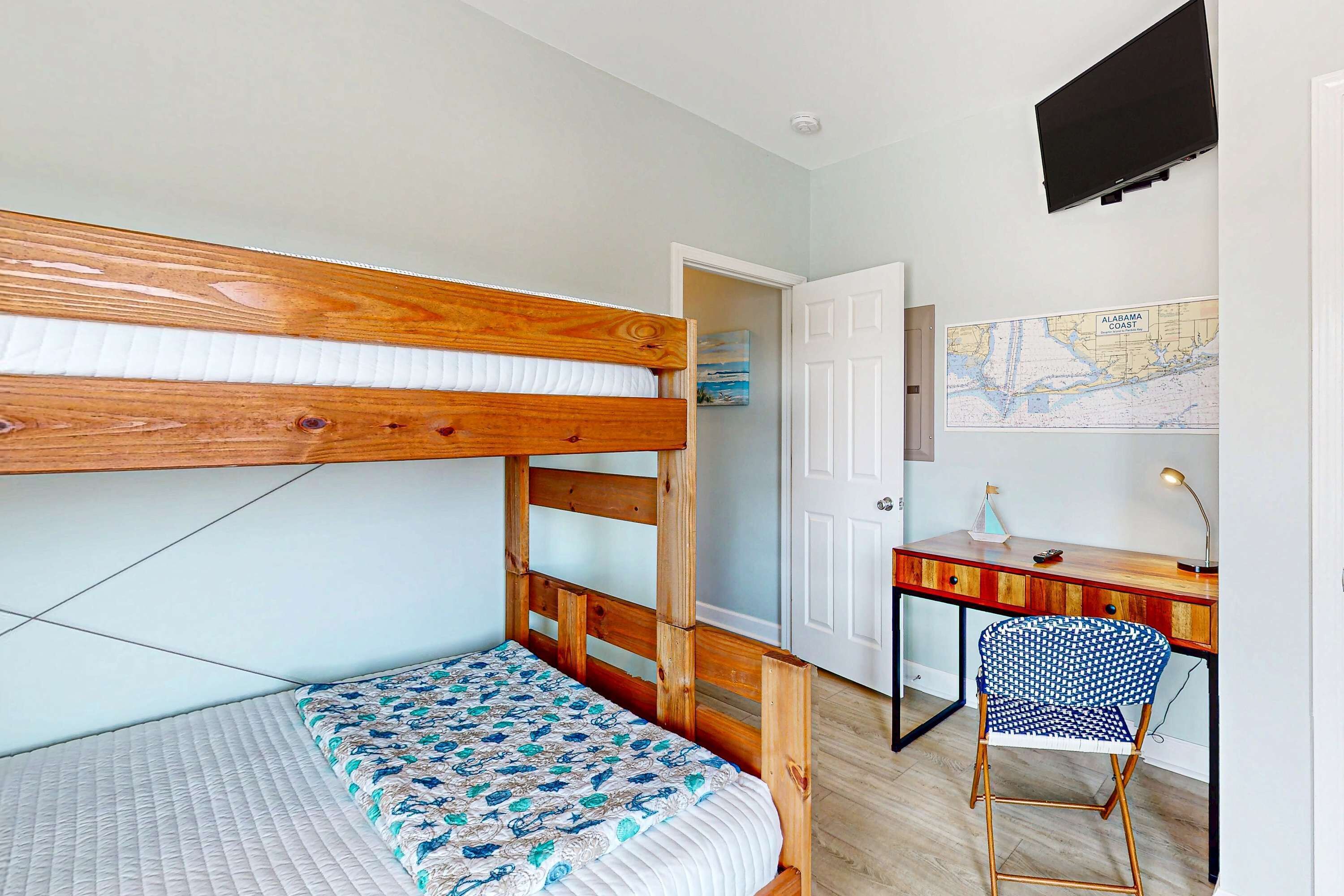 A bunk bed with a patterned bedspread, a desk with a chair, a map on the wall, and a television mounted above the desk.