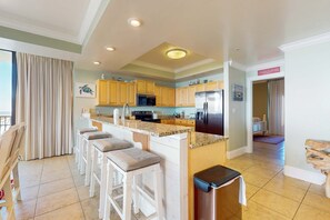 A modern kitchen with a central island, bar stools, wooden cabinets, and a stainless steel refrigerator. - Beachfront condo with indoor & outdoor pools, hot tub, fitness center, tennis (Dauphin Island)