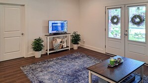 Una habitación con una televisión sobre un mueble bajo, una mesita con un adorno, un tapete azul con diseño y dos macetas con plantas.