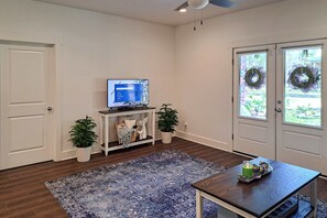 A room with a television on a stand, a small table with a decorative item, a blue patterned rug, and two potted plants. - Cozy cottage located near the beach with gas grill, yard, & washer & dryer (Dauphin Island)