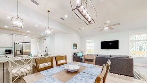 Eine moderne Küche mit weißen Schränken, ein Essbereich mit Tisch und Stühlen, ein graues Sofa und ein Deckenventilator.