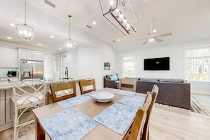 A modern kitchen with white cabinets, a dining area with a table and chairs, a gray sofa, and a ceiling fan.