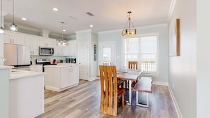 A modern kitchen with white cabinetry, a dining area with a wooden chair, and a hanging light fixture. - Exquisite oceanfront beach home with hot tub, central AC, balcony, & W&D (Dauphin Island)