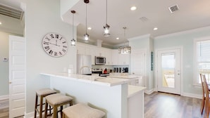A modern kitchen with a central island, bar stools, pendant lights, and a large wall clock. - Exquisite oceanfront beach home with hot tub, central AC, balcony, & W&D (Dauphin Island)