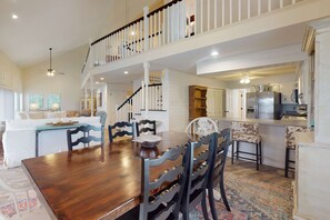 A spacious dining area with a wooden table and chairs, a staircase leading to an upper level, and a kitchen visible in the background. - Bayfront home with boat dock, water views, deck, & fish cleaning station (Dauphin Island)