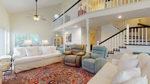 Ein geräumiges Wohnzimmer mit einem großen, gemusterten Teppich, einem weißen Sofa, einem Relaxsessel und einem blauen Sessel. Eine Deckenlampe und eine Treppe mit weißen Geländern sind zu sehen.